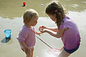3 year old girl and 18 month old baby girl, on the beach looking at a crab that they ve found.