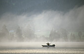 Fisher in a boat on Kochelsee lake in morning mist, Bavaria, Germany