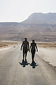 A young man and woman covered in mud walking to the Dead Sea, desert scenery behind them, Israel.
