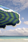 Striped fringed beach umbrella in left of frame, sea and blue sky with puffy clouds in right and top.