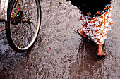 Barefooted woman in a skirt walking on wet muddy ground next to a rickshaw wheel