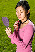 Young Spanish/Asian woman putting on makeup at a park.