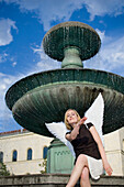 Mid adult woman wearing angel wings sitting near fountain at university, Munich, Bavaria, Germany
