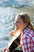 A young woman sitting on the banks of the river Isar in the evening playing the guitar, Munich, Bavaria, Germany