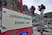 Schild Europäischer Fernwanderweg E5 mit zwei Frauen außerhalb des Schärfebereichs unterwegs auf Wanderweg, nahe Memmingher Hütte, Lechtaler Alpen, Tirol, Österreich