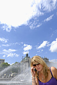 Young woman phoning with mobile phone near a fountain at Stachus, Munich, Bavaria, Germany