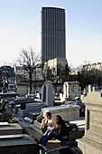 Two female students discuss their homework in Cimetiere du Montparnasse with the high-rise Tour Montparnasse in the background. Paris. France