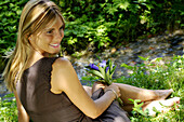 Frau sitzt auf eine Wiese in der Nähe von einem Bach mit einer Blume in der Hand und entspannt sich, Urlaub, Südtirol, Italien