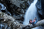Zwei Paare sitzen auf einem Baumstamm vor einem Wasserfall, See, Tirol, Österreich