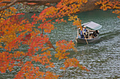 A pleasure boat framed in red-ornge maple leaves being poled along the pond in Arashiyama