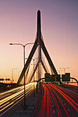 Rush hour traffic light-trails stream in and out of the city of Boston over the Leonard P Kakim Bunker Hill Bridge,  with a colorful sky in the background. Massachusetts,  USA