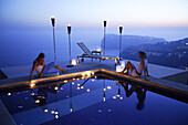 Two young women sitting by the side of a pool with candles and torches, Cala Llamp, Andratx, Mallorca, Balearic Islands, Spain