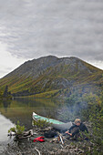 Mann sitzt am Lagerfeuer am Ufer des St. Elias Sees, Kluane-Nationalpark, Yukon, Kanada