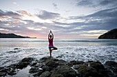 Young woman doing yoga on rocks at sunset in front of ocean surf at Playas del Coco, Costa Rica