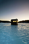 An ice-fishing shanty sitting on a frozen lake is sihouetted against the fading light on a winter evening