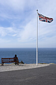 Frau auf Bank und wehende Union Jack Flagge an Fahnenmast, Land's End, Cornwall, England, Europe