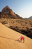 Woman climbing at red rock face, Great Spitzkoppe in background, Great Spitzkoppe, Namibia
