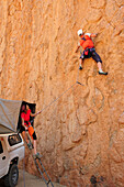 Man climbing at red rock face, woman sitting in roof tent belaying, Great Spitzkoppe, Namibia