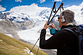 Hiker with headphones photographing (myclimate audio trail) near Gorner glacier, Monte Rosa and Liskamm in background, Zermatt, Canton of Valais, Switzerland