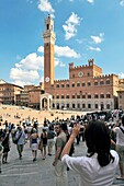 Tourists in the Piazza del Campo, the central square of the city of Sienna, Tuscany, Italy Torre del Mangia tower rises behind