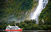 New Zealand, Southland, Fiordland National Park A red tour boat neat a waterfall cascading through a Beech forest on the edge of the Milford Sound
