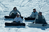 Four young snowboarders sitting on ski slope, looking away