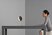 Frau sitzt am Tisch, Gesicht im Spiegel reflektiert
