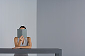Woman sitting at table reading, face obscured by book