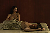 Nude couple in bed, man lying on back, woman sitting up, both looking away