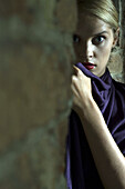 Woman hiding behind brick wall, staring at camera with wide eyes
