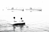 People on water in inflatable raft with pedal boats in background, b&w