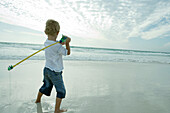 Boy holding fishing pole over shoulder on beach, rear view