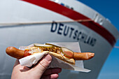 Hand hält Polser Hot Dog: Passagiere von Kreuzfahrtschiff MS Deutschland (das ZDF Traumschiff der Reederei Peter Deilmann) werden bei Ankunft mit einer Wurst gratis begrüßt, Aalborg, Nordjütland, Dänemark
