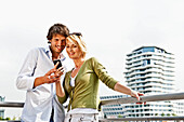 Lächelndes Paar mit einem Handy lehnt an einem Geländer, Marco-Polo-Tower im Hintergrund, HafenCity, Hamburg, Deutschland