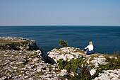 Woman sits on cliff at Högklint Naturreservat and admires coastline, Visby, Gotland, Sweden