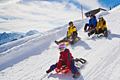 Ein Mädchen, eine Frau und zwei Männer beim Schlittenfahren auf der Schlittelbahn im Wintersportgebiet Stoos mit dem modernen Schlitteln Scorpion Racer, Kanton Schwyz, Schweiz
