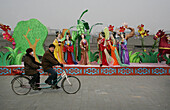 A Chinese couple riding a tandem bicycle past a traditional display, near the Old City wall, Xian, Xi'an, Capital of Shaanxi Province, China
