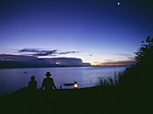 Fisherman in dugout canoe going past as couple having sundowner drinks at sunset on Domwe Island, Lake Malawi, Malawi