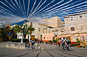 Cyclists going past square with white bunting in Caimari, Majorca, Spain
