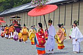 Costumed participants in the parade, some carrying red parasols for ladies walking beside them with the Imperial Palace in the background