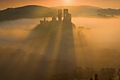 Corfe Castle back lit by the sun causing long shadows to appear as it rises out of the mist at dawn Purbeck Dorset
