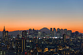 A telephoto view in clear winter twilight captures Mt. Fuji's distinctive peak rising dramatically beyond the skyscrapers (including Tokyo City Hall) and the city lights of the Shinjuku District in Tokyo, Japan.