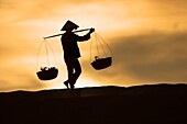 Woman with conical hat carries pannier baskets in sunrise along crest of sand dunes near Mui Ne Vietnam