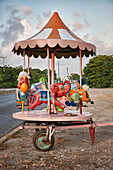 Childs merry go round next to a road. Small cart with platform and carousel, with seats.
