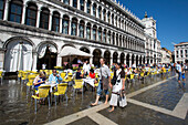 Junges Paar läuft barfuß über den überfluteten Markusplatz während Menschen vor einem Café sitzen, Venedig, Venetien, Italien, Europa