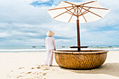 Frau mit vietnamesischer Kleidung und Strohhut blickt auf das Meer mit Fischerbooten, Strand von Mui Ne, Südvietnam, Vietnam, Asien