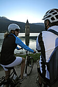 Couple on electric bikes at Lake Reschensee, Church tower in the background, E-bikes, Bozen, South Tyrol, Italy