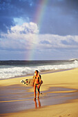 Sexy girl in string bikini walking along the beach holding surfboard, rainbow behind.