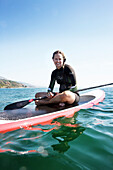 USA, California, Malibu, El Pescador Beach, an athletic woman sits on her paddleboard and paddles through the water