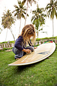 INDONESIA, Mentawai Islands, Kandui Surf Resort, girl waxing surfboard on lawn with palm trees in the background